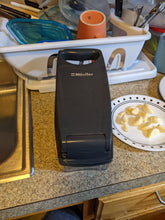 Load image into Gallery viewer, View of the top of the device with the lid closed. It is all gray with &quot;Müeller&quot; on top and a handle on one end. Sitting beside it are garlic cloves on a plate, waiting to be chopped.
