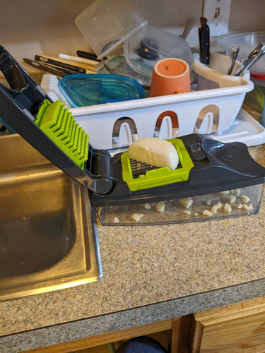 Chopper device with clear base, gray cover and lime green accents around the blades sits on a kitchen counter with a big chunk of onion on it, waiting to be chopped.