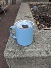 Load image into Gallery viewer, Blue mug with clear lid and black tab that opens the hole from which to drink. The mug is on a concrete planter wall.
