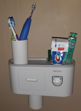 Load image into Gallery viewer, The full toothbrush holder and dispenser is shown hanging on the wall. Toothbrushes sit in a cup on top, and two tubes of toothpaste and deodorant are on the other side. A cup hangs from below.
