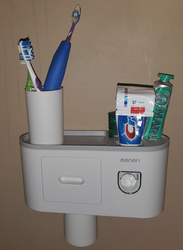 The full toothbrush holder and dispenser is shown hanging on the wall. Toothbrushes sit in a cup on top, and two tubes of toothpaste and deodorant are on the other side. A cup hangs from below.