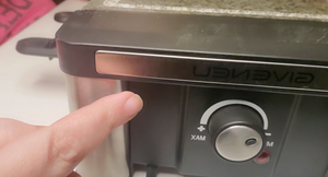A close up image of a hand pointing to the dial that controls the heat on the grill. It is one knob that turns from M (low) to XAM (high). There is a light beside the knob. 
