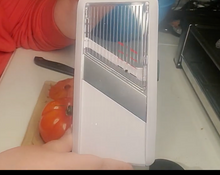 Load image into Gallery viewer, Surface of the mandoline slicer. the Top part is clear, the middle blade is gray, and the bottom is white. The frame is white. A tomato and arm of a person are visible behind the mandoline slicer. 
