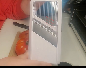 Surface of the mandoline slicer. the Top part is clear, the middle blade is gray, and the bottom is white. The frame is white. A tomato and arm of a person are visible behind the mandoline slicer. 