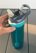 Load image into Gallery viewer, The bottle has a turquoise, see-through base with a gray lid with the spout open. The spout has a clear, thick cover over it. There is a fixed, plastic two-finger &quot;loop&quot; that is part of the lid. A white button is visible on the front, used to release the spout. A small slide button is visible, used to lock the spout. The bottle is sitting on the ground with a person&#39;s hand holding it.
