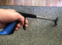 Load image into Gallery viewer, A hand is wrapped around the reacher-grabber handle, with their fingers on the lever to operate the device. The handle is blue and black and then the length of the device is silver, with a black grabber on the end. It is shown reaching toward the floor, which is a gray carpet.
