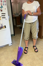 Load image into Gallery viewer, A person standing in a kitchen holding the Swiffer mop, which has a purple handle, a silver rod extension, and then a purple base that holds the cleaning fluid and the swivel mop head.
