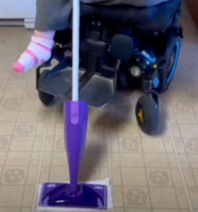 The purple Swiffer WetJet head is visible on the floor. A power wheelchair is in the background.