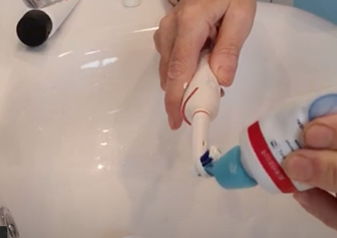 A person is using a toothpaste tube with the self closing lid to dispense toothpaste onto an electric toothbrush over a white sink.