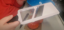 Load image into Gallery viewer, White OXO mandoline slicer with silver blade, clear adjustment piece, and a black handle. A person is holding the slicer, and a tomato and knife are visible on the table.
