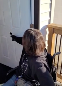 A woman with brown hair, who is a wheelchair user, is using the black door closer to close the front door to a house.