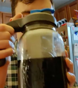Side view of mason jar filled half way with coffee. A person is holding it up by the gray handle attached to the lid. 