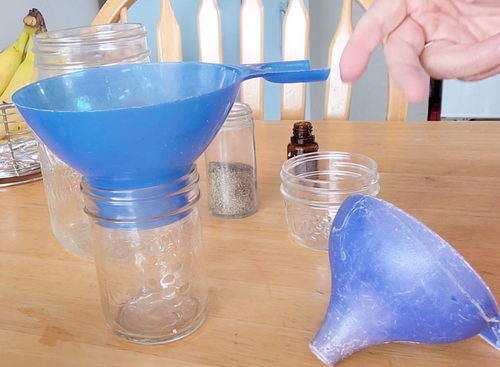 Two blue funnels on a table. One is sitting in a Mason jar and the other is sitting on the table. Other jars are visible on the table.
