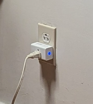 A close-up of the smart plug plugged into a wall. A blue light glows on one side. A cord is plugged into the smart plug.