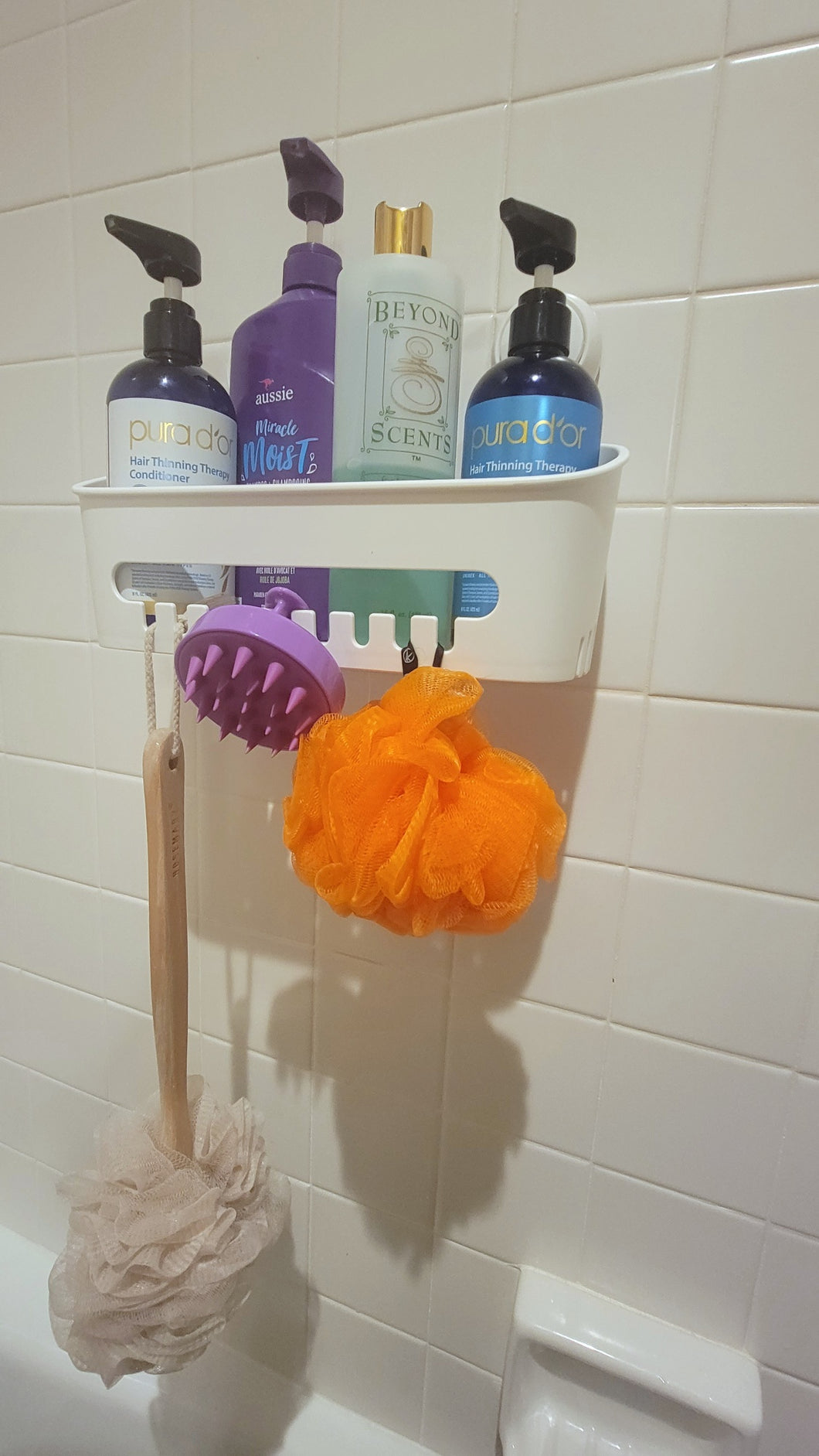 White shower caddy attached to a white tile shower wall and filled with four different bottles of soaps/shampoos. Hanging from the caddy is a scalp brush, long-handle loofah, and an orange loofah.