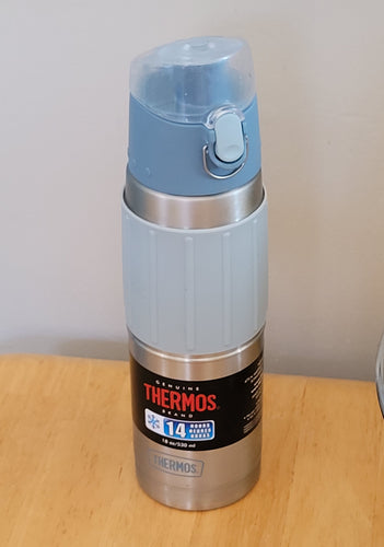 Thermos water bottle on a wood table. It is light blue on top with a stainless steel container and a light blue silicone sleeve with ridges. At the bottom is a black label that says 