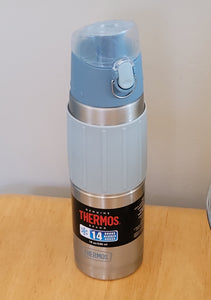 Thermos water bottle on a wood table. It is light blue on top with a stainless steel container and a light blue silicone sleeve with ridges. At the bottom is a black label that says "Genuine Thermos Brand" and 14. 