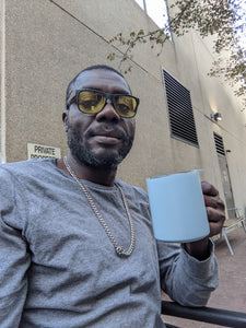 Tony, a black man with a trimmed beard, glasses, a gray shirt and a silver chain, is holding the blue MiiR mug.