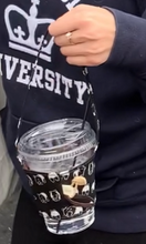 Load image into Gallery viewer, A person is using the Uma hana black and white drink carrier to carry water in a plastic cup with a lid and straw.
