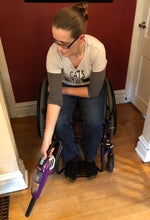 Load image into Gallery viewer, A white woman with brown seated in a wheelchair is using the Bissell Pet Hair Eraser Cordless Hand Vacuum with the crevice tool extension to vacuum her floor.
