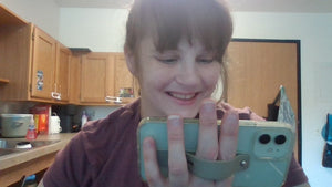 A white woman with brown bangs and hair pulled back is looking at her phone and smiling. Her phone is held in landscape mode, with two fingers in the strap, supporting the phone.