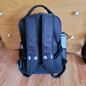 View of the bag from the back. Two "shoulder" straps (like a backpack) are visible, each with a small reflector strip across the middle of the strap. The handle is clearly visible on top.