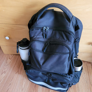 The backpack is sitting on the ground. A large water bottle and a travel coffee mug are in the two side pockets on the bag. A silver reflector strip is visible across the front pocket.
