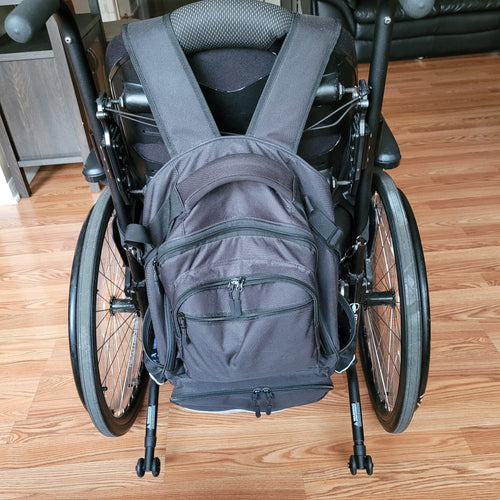 Black wheelchair bag hangs by two straps on the back of a wheelchair. The bag features several pockets with zippers and elastic pouches (for example, for bottles) on either side. There is a handle on top of the bag.