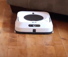 Load image into Gallery viewer, White, square shaped robot device with black features on a hardwood floor.
