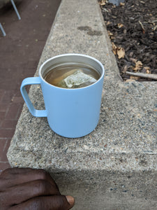Light blue mug with a tea bag and tea in it, sitting on a concrete planter wall.