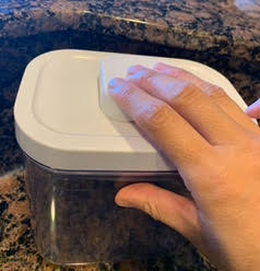 Pop container, clear with a white lid, sitting on a marble countertop. An adult hand is pushing the button on the lid.