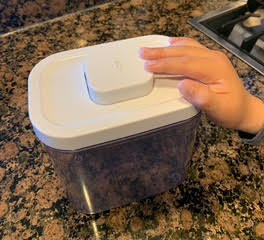 Pop container, clear with a white lid, sitting on a marble countertop. A child's hand is pushing the button on the lid.