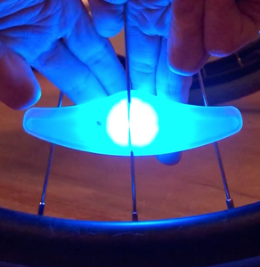 Spoke light lit up in blue. It is attached to the spokes of a wheel and a person is pushing a button on the back using several fingers.